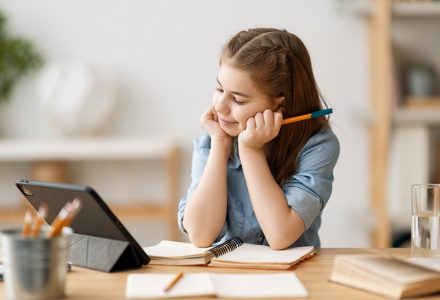 An image of a girl who is looking at a tablet, and holding a pencil, taking lessons online.
